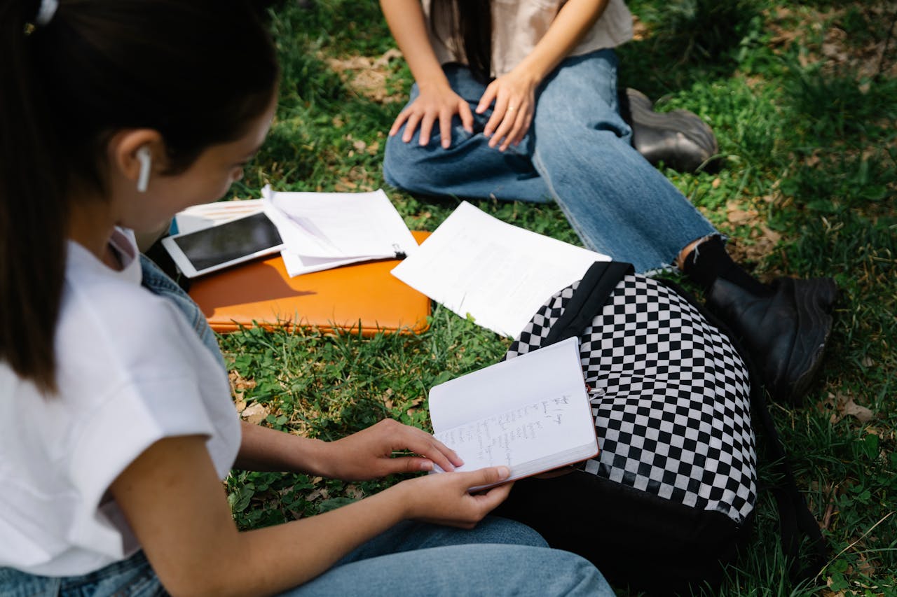 Two students studying together outdoors on a sunny day, surrounded by grass and books.